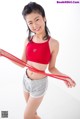 A woman in a red sports bra top and gray shorts holding a red resistance band.